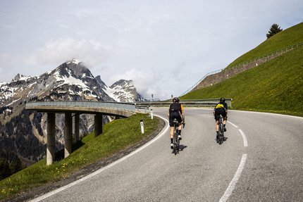 Two cyclists riding on a mountain road with snow-capped peaks in the background