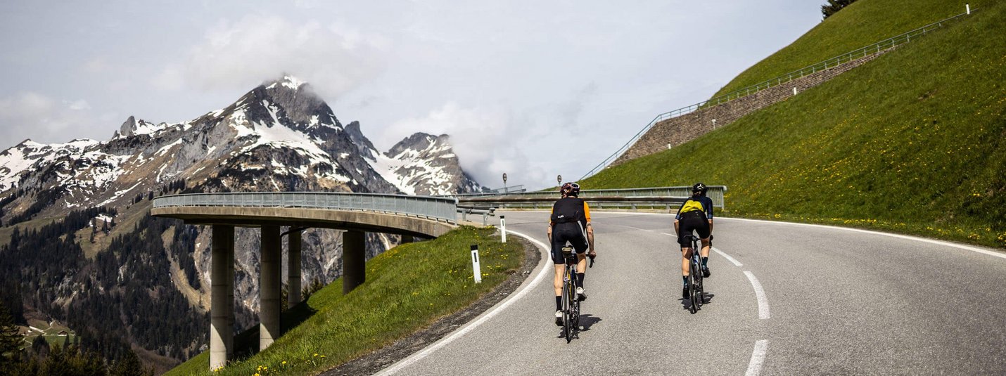 Two cyclists riding on a mountain road with snow-capped peaks in the background