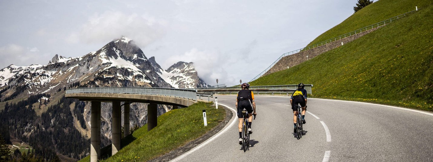 Zwei Radfahrer fahren eine Bergstraße mit schneebedeckten Gipfeln im Hintergrund
