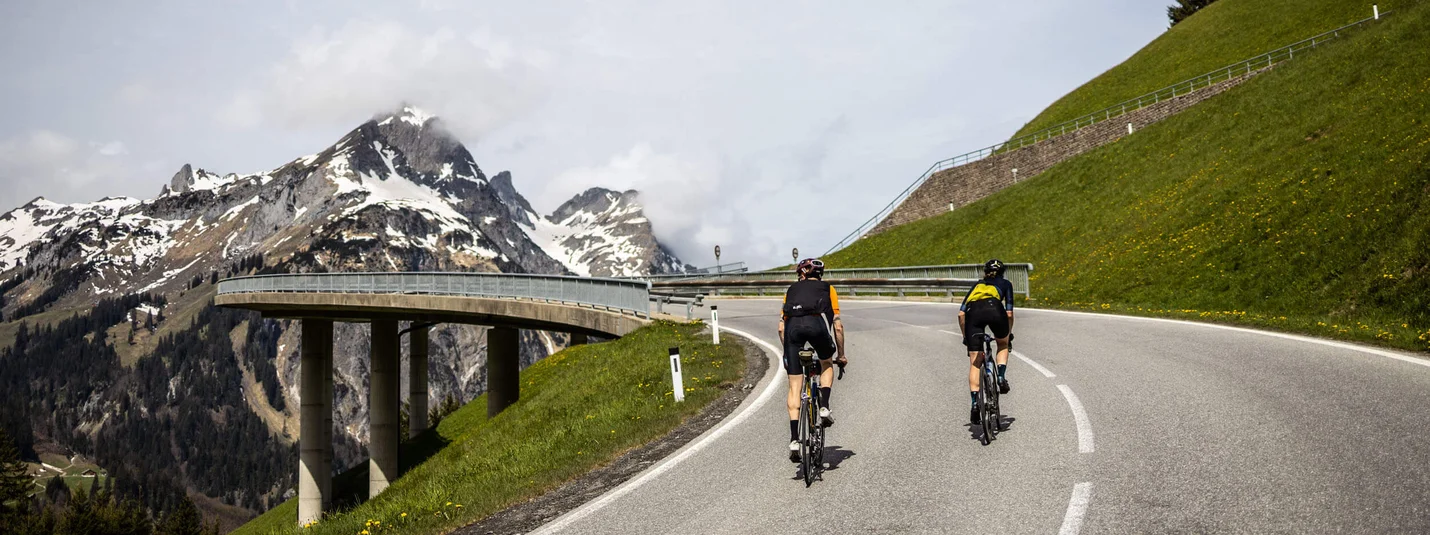 Racefietsvakantie in Vorarlberg © Bregenzerwald Tourismus - Merlijn Spenkerlink Twee fietsers op een bergweg met besneeuwde toppen op de achtergrond
