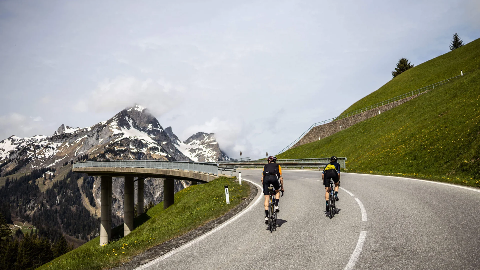 Zwei Radfahrer fahren eine Bergstraße mit schneebedeckten Gipfeln im Hintergrund