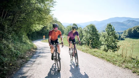 Two cyclists riding on a country road with wooded hills in the background