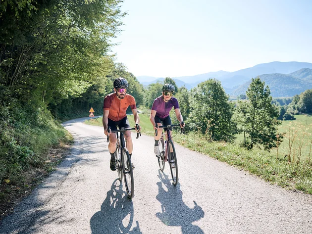 Two cyclists riding on a country road with wooded hills in the background