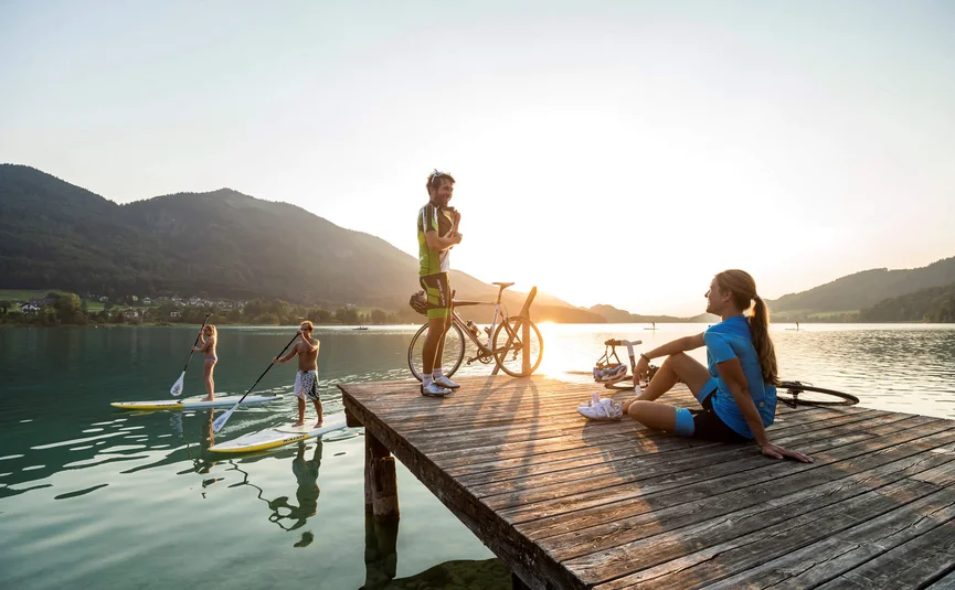 Personen auf Steg und Stand-up-Paddle-Boards bei Sonnenuntergang am See