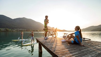 Personen auf Steg und Stand-up-Paddle-Boards bei Sonnenuntergang am See
