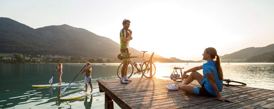 Personen auf Steg und Stand-up-Paddle-Boards bei Sonnenuntergang am See