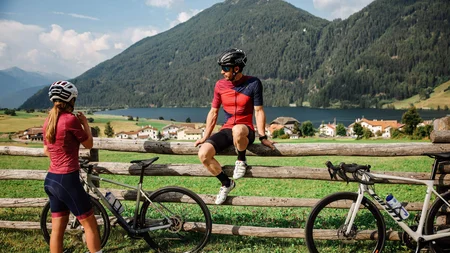 Zwei Radfahrer machen Pause an einem Zaun mit Berglandschaft im Hintergrund