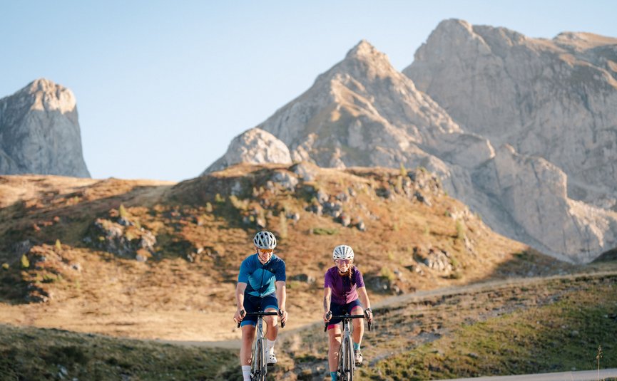 Two cyclists riding on mountain road with rocky peaks under clear sky