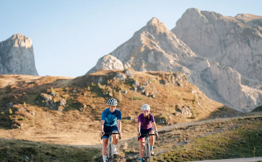 Due ciclisti su strada di montagna con picchi rocciosi sotto cielo sereno