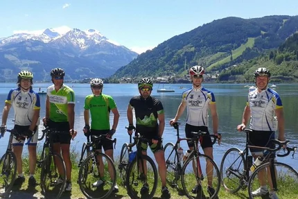 Zes wielrenners met racefietsen bij een bergmeer met besneeuwde toppen