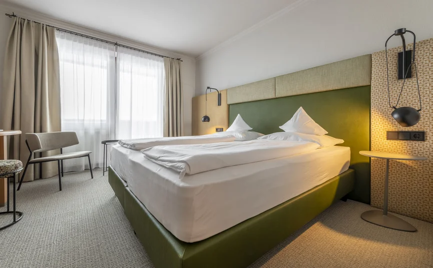 Camera d'albergo luminosa con letto verde e due lampade sospese accanto al tavolino