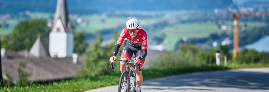 Der Kufsteinerland Radmarathon © Kufsteinerland / Martin Bihounek Radfahrer in rotem Trikot fährt auf Landstraße mit Dorf und Bergen im Hintergrund