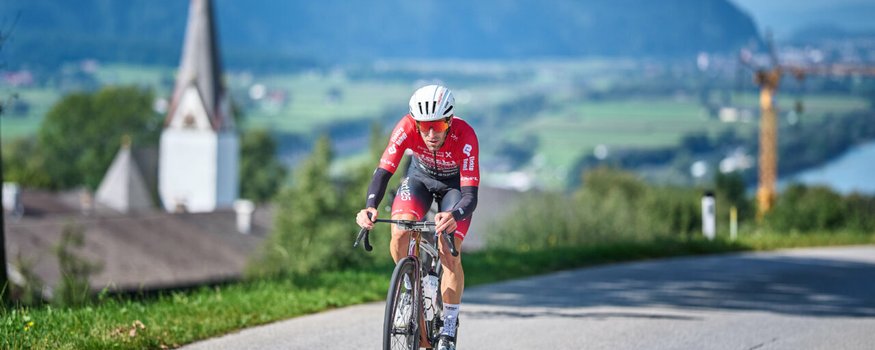 Der Kufsteinerland Radmarathon © Kufsteinerland / Martin Bihounek Radfahrer in rotem Trikot fährt auf Landstraße mit Dorf und Bergen im Hintergrund