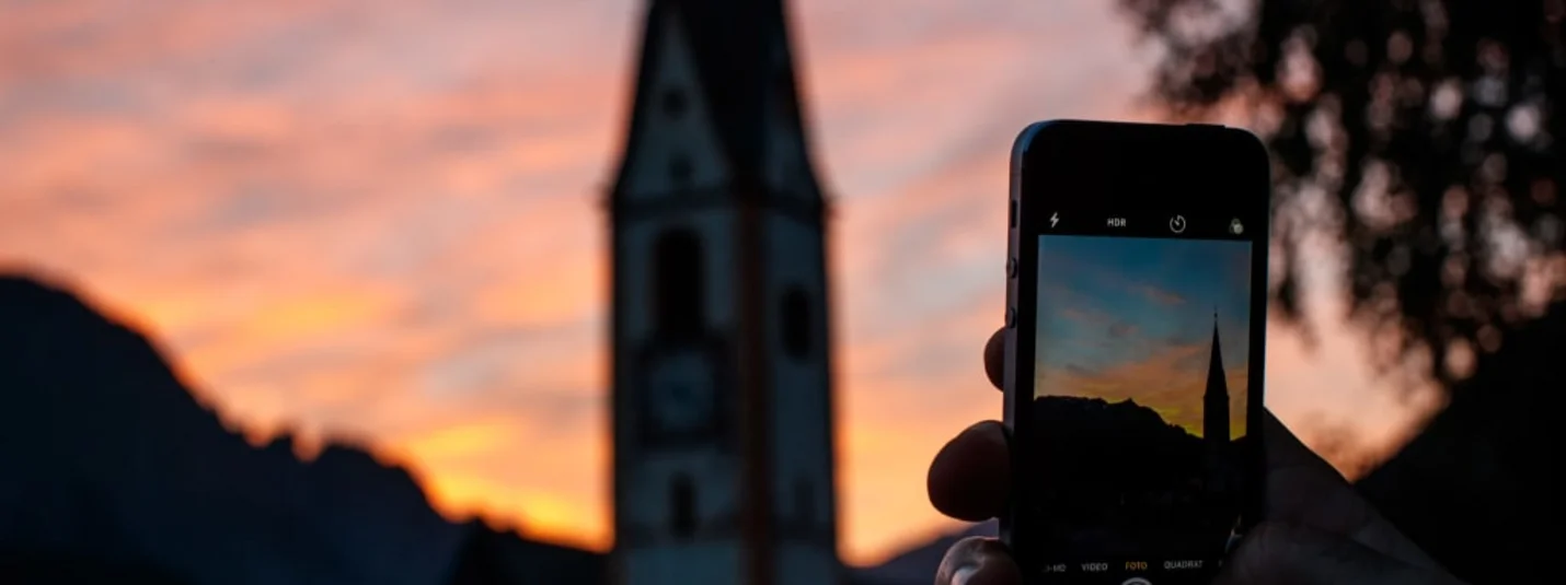 7- Tage Wanderpackage Hand hält Smartphone und fotografiert Kirchturm bei Sonnenuntergang