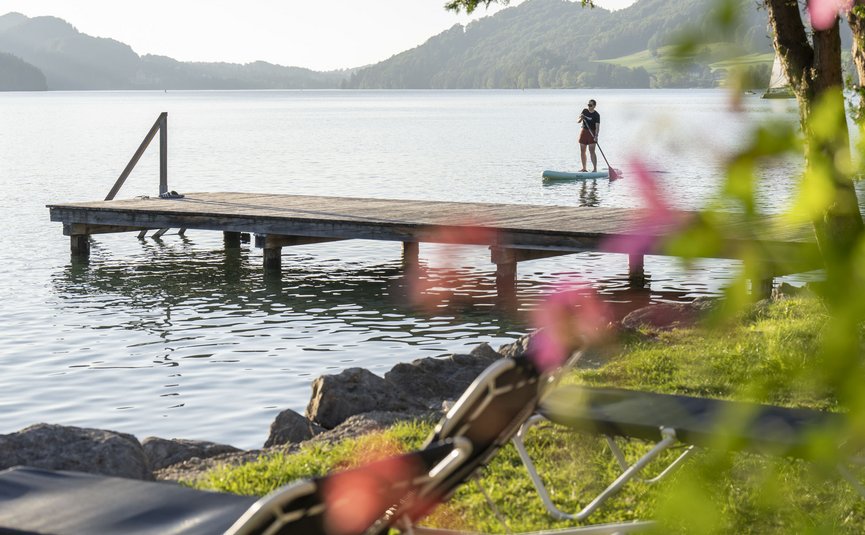 Person paddleboarding near wooden dock on lake with forested hills