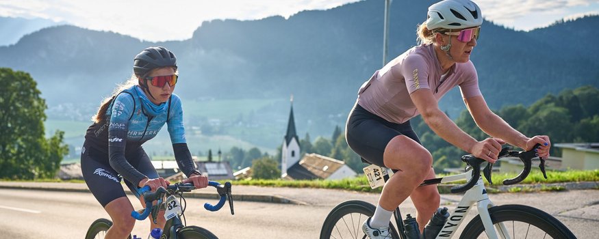Der Kufsteinerland Radmarathon © Kufsteinerland / Martin Bihounek Zwei Rennradfahrerinnen mit Helm fahren auf einer Straße bei Bergpanorama