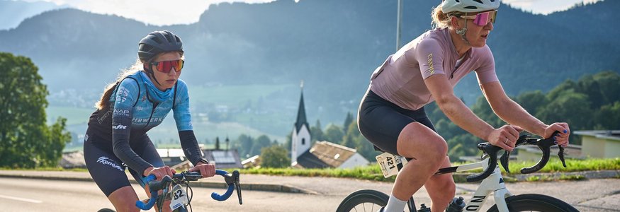 Der Kufsteinerland Radmarathon © Kufsteinerland / Martin Bihounek Zwei Rennradfahrerinnen mit Helm fahren auf einer Straße bei Bergpanorama
