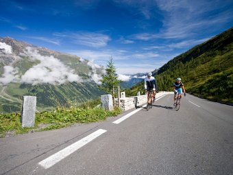 Zwei Radfahrer fahren auf einer Bergstraße mit Bergen und blauem Himmel im Hintergrund