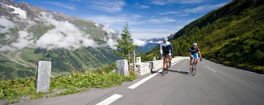 Zwei Radfahrer fahren auf einer Bergstraße mit Bergen und blauem Himmel im Hintergrund