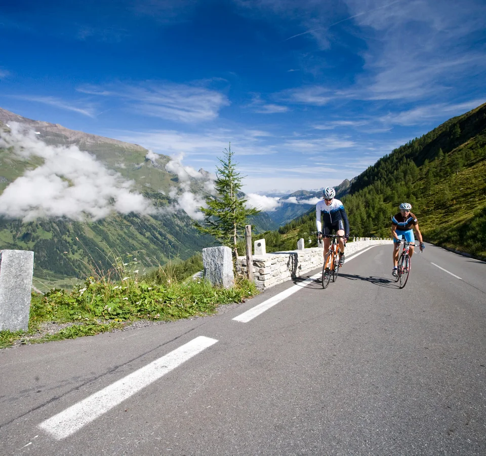 Zwei Radfahrer fahren auf einer Bergstraße mit Bergen und blauem Himmel im Hintergrund