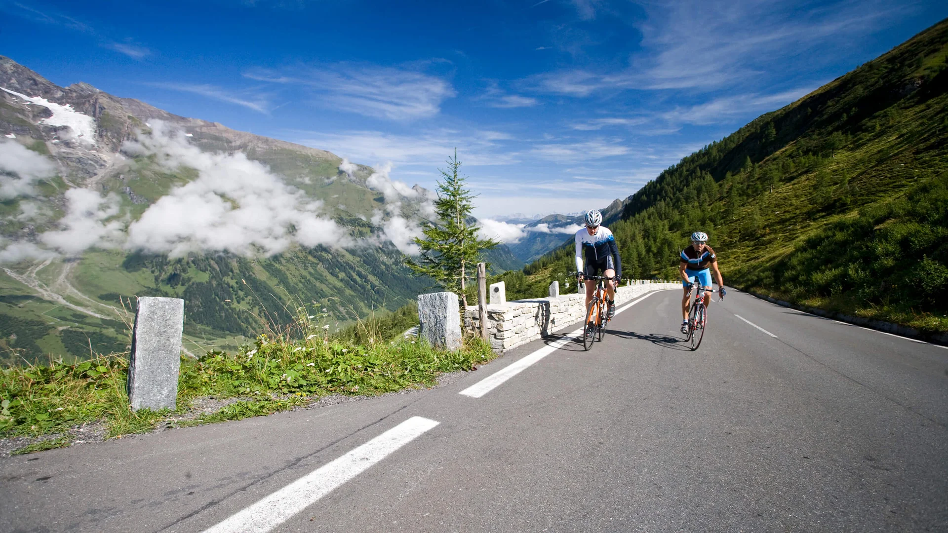 Zwei Radfahrer fahren auf einer Bergstraße mit Bergen und blauem Himmel im Hintergrund