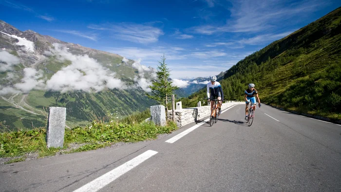 Zwei Radfahrer fahren auf einer Bergstraße mit Bergen und blauem Himmel im Hintergrund