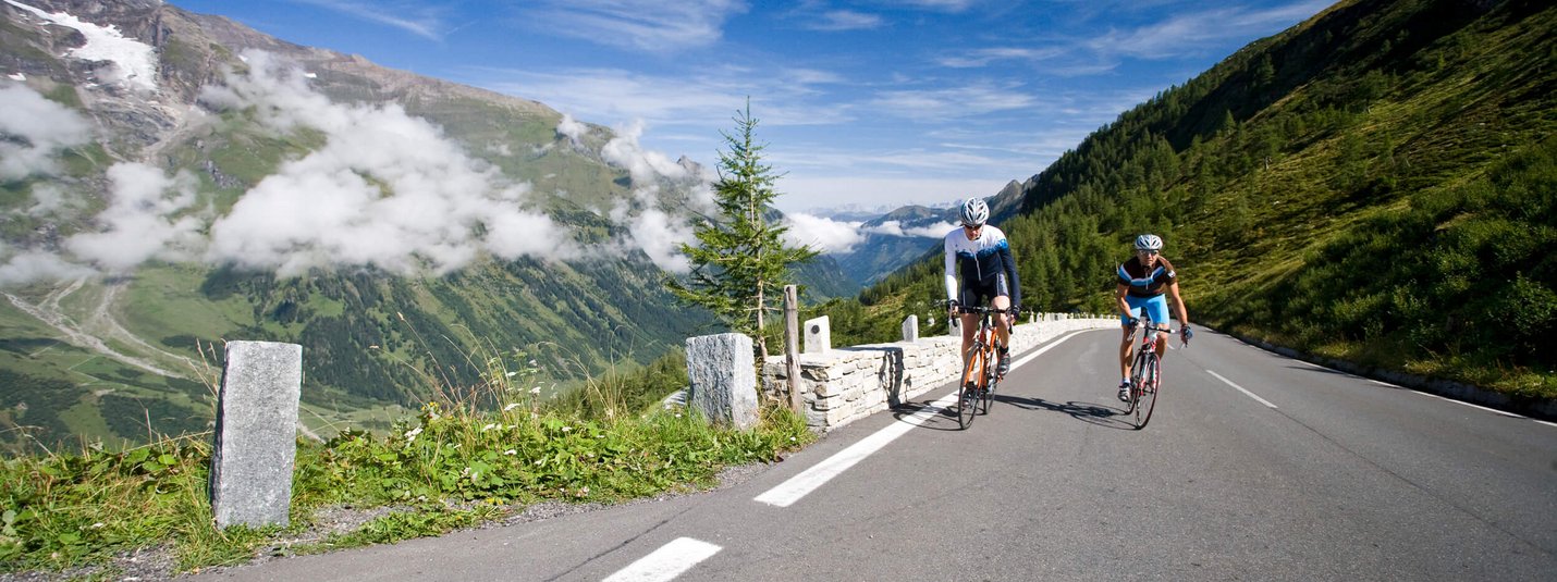 Due ciclisti su una strada di montagna con montagne e cielo azzurro sullo sfondo