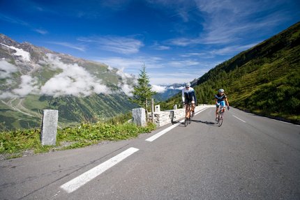 Due ciclisti su una strada di montagna con montagne e cielo azzurro sullo sfondo