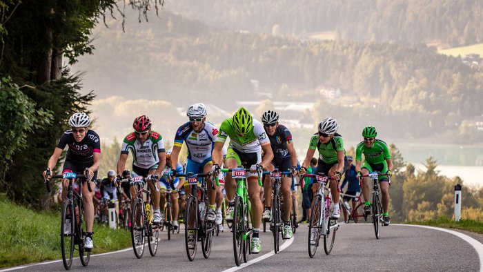 Ciclisti in gara su strada collinare con foresta e lago sullo sfondo