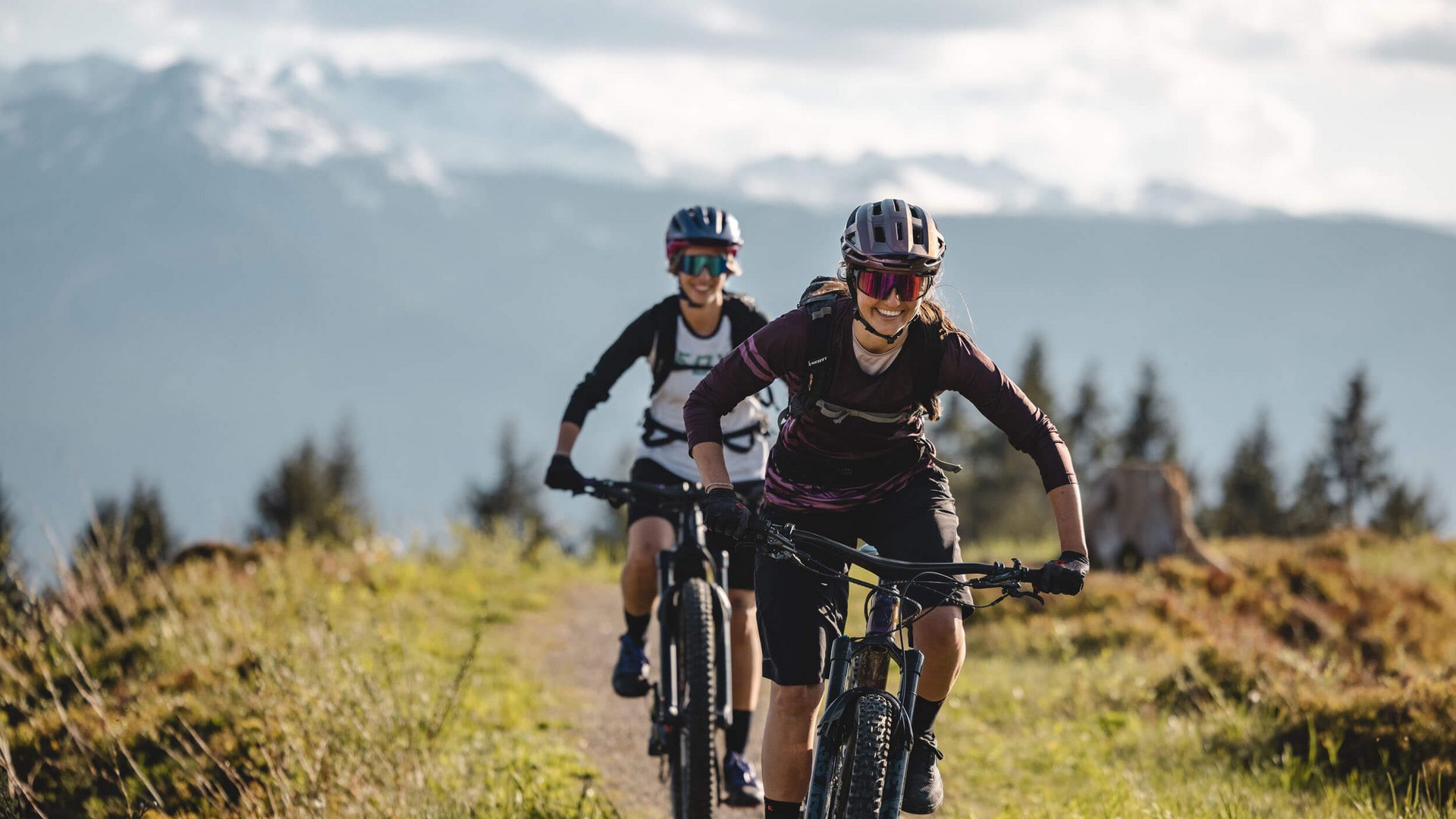 Zwei Frauen fahren Mountainbike auf Bergpfad mit Bergen im Hintergrund