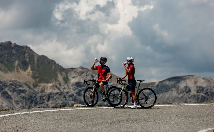 Two cyclists drinking water on a mountain road under cloudy sky