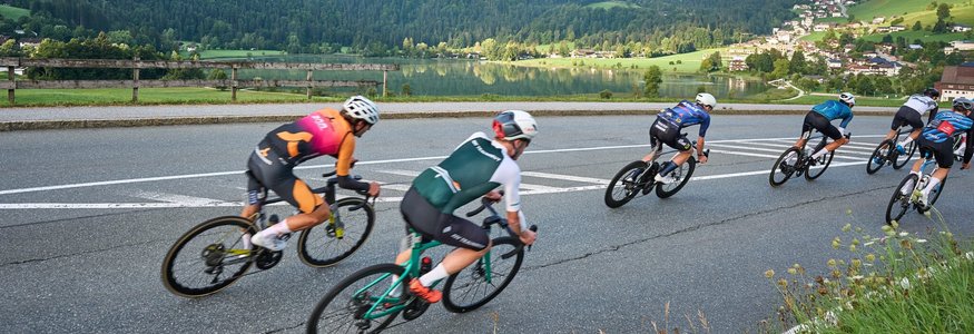 Der Kufsteinerland Radmarathon © Kufsteinerland / Martin Bihounek Radrennfahrer fahren bergab auf kurviger Straße mit Bergen und See im Hintergrund