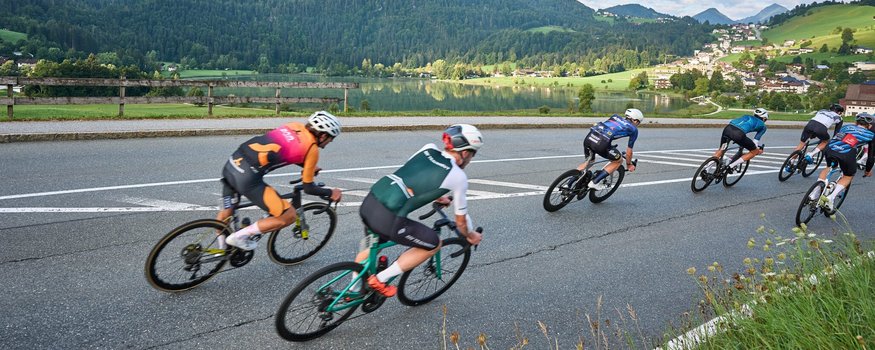 Der Kufsteinerland Radmarathon © Kufsteinerland / Martin Bihounek Radrennfahrer fahren bergab auf kurviger Straße mit Bergen und See im Hintergrund