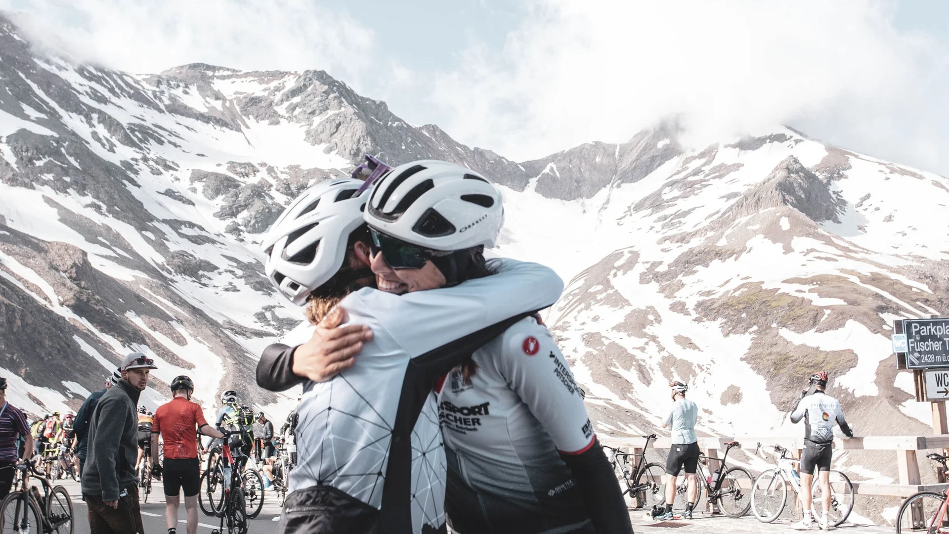 Two cyclists hugging with snow-covered mountains in the background at cycling event
