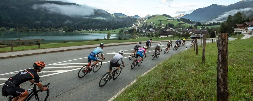 Gruppo di ciclisti su strada con montagne e lago sullo sfondo