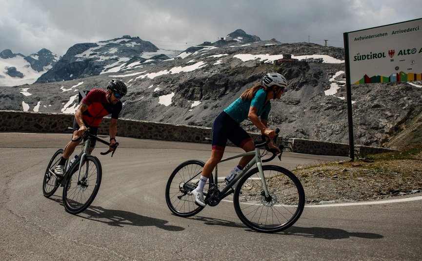 Twee fietsers op bergweg met besneeuwde toppen en Zuid-Tirol bord