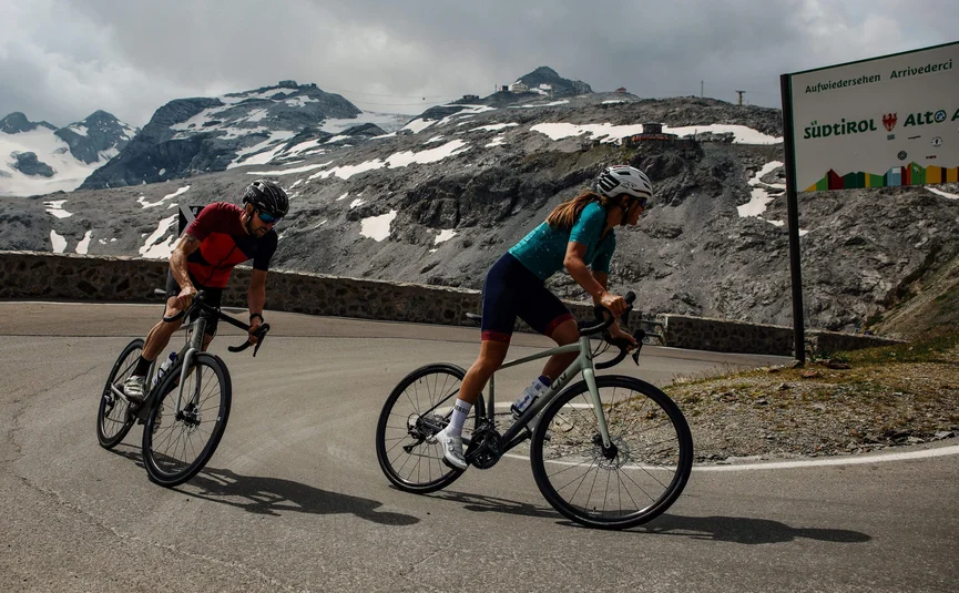 Two cyclists riding mountain road with snowy peaks and South Tyrol sign