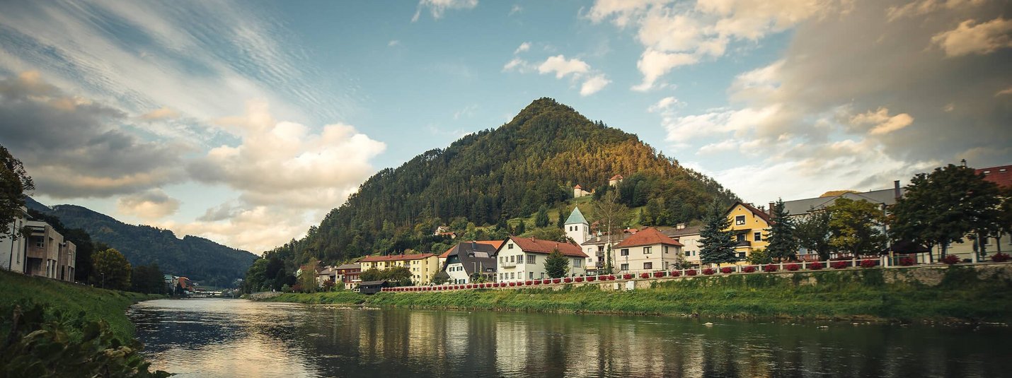 Riverbank with houses and wooded hill under cloudy sky
