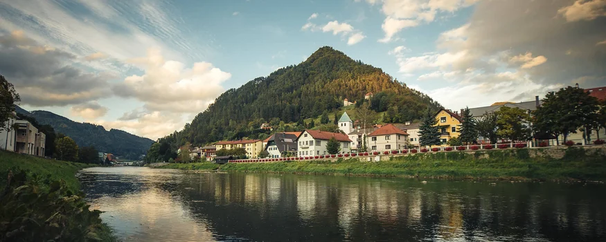 Savinja Tal © Jacob Riglin - www.slovenia.info Flussufer mit Häusern und bewaldetem Hügel unter bewölktem Himmel