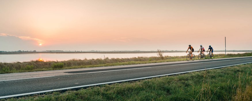 Three cyclists riding by a river at sunset