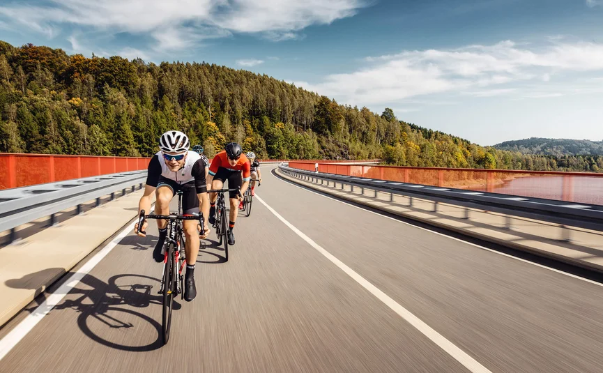 Radfahrergruppe auf Straßenbrücke mit Wald im Hintergrund bei sonnigem Wetter