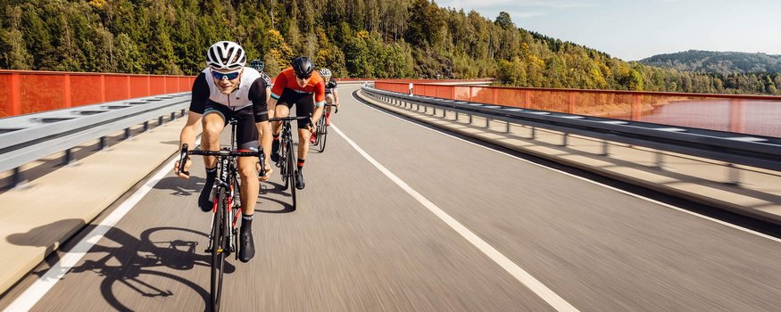 Radfahrergruppe auf Straßenbrücke mit Wald im Hintergrund bei sonnigem Wetter