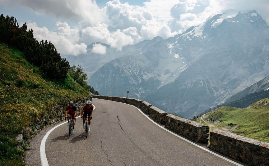 Two cyclists on mountain road with snowy peaks in background