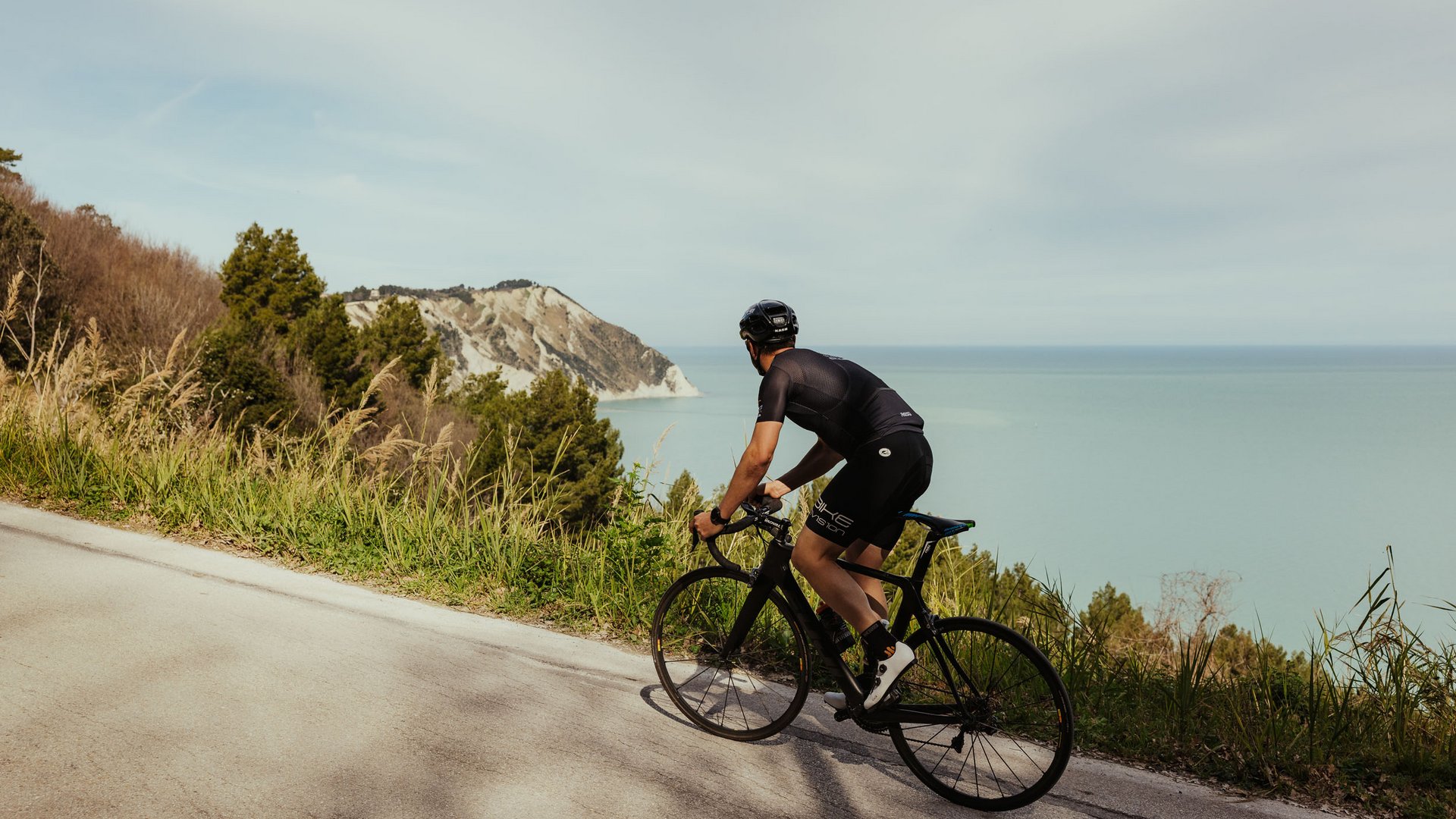 Radfahrer auf Straße mit Meerblick und Hügel im Hintergrund