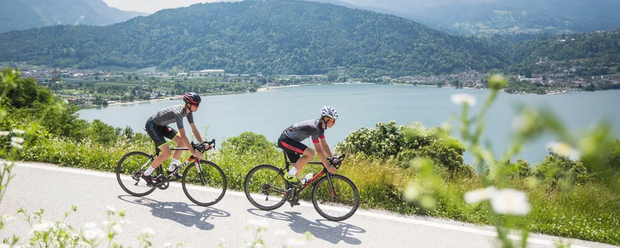 Levico See © Heiko Mandl Zwei Radfahrer fahren an einem See vor bewaldetem Bergpanorama