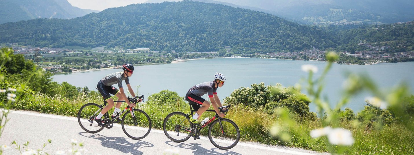 Two cyclists riding beside a lake with green mountains in the background