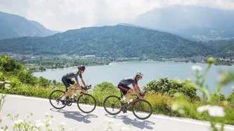 Due ciclisti pedalano vicino a un lago con montagne verdi sullo sfondo