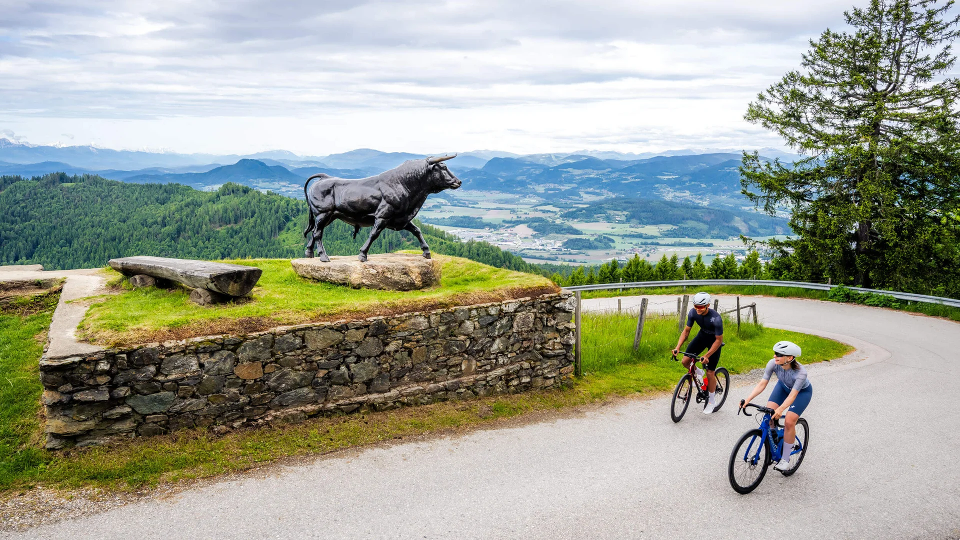 Two cyclists riding past a bull statue on a mountain road
