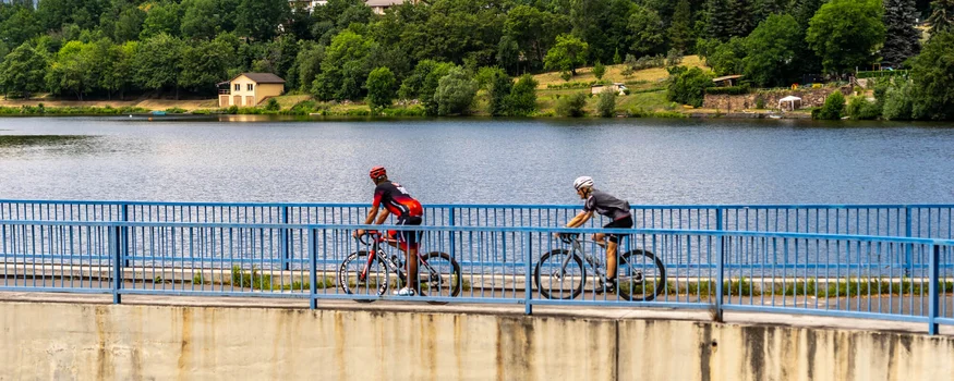 Zwei Radfahrer auf einer Brücke über einem See, Häuser im Hintergrund
