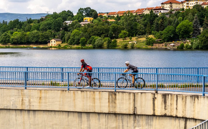 Due ciclisti su un ponte sopra un lago, case e alberi sullo sfondo